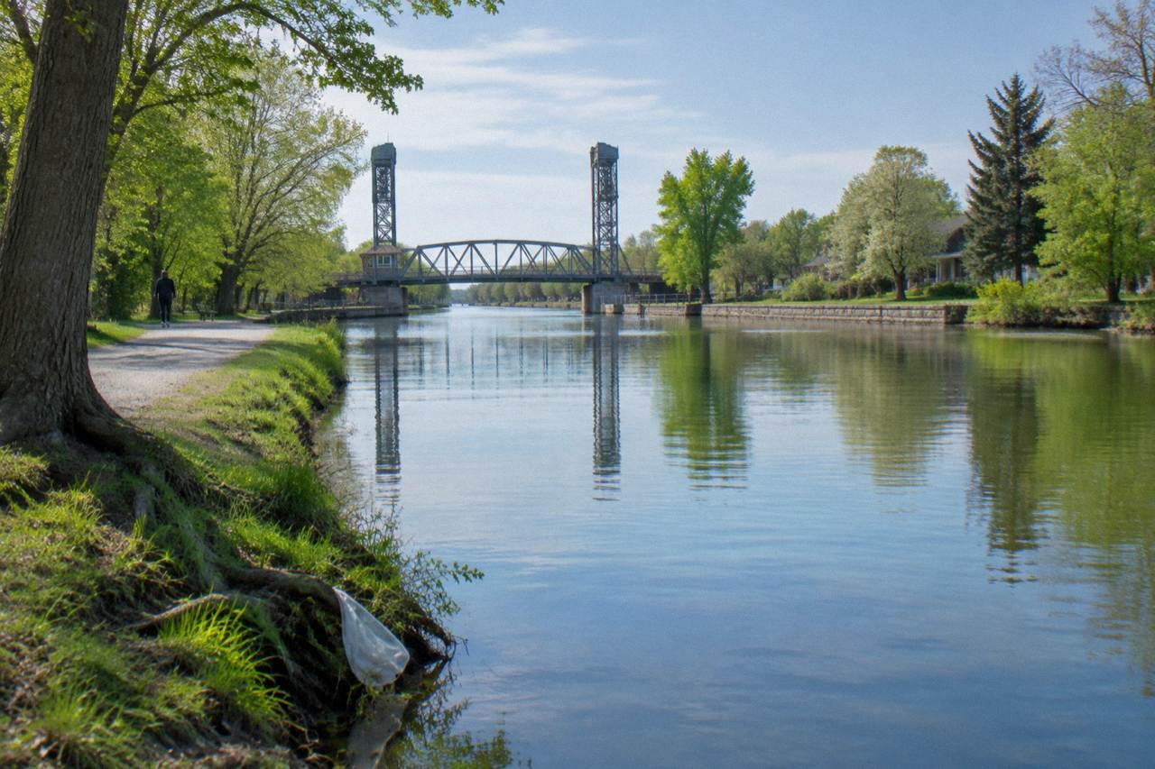 A wide calm stretch of the Erie Canal reflecting trees and an empty towpath under a bright sky