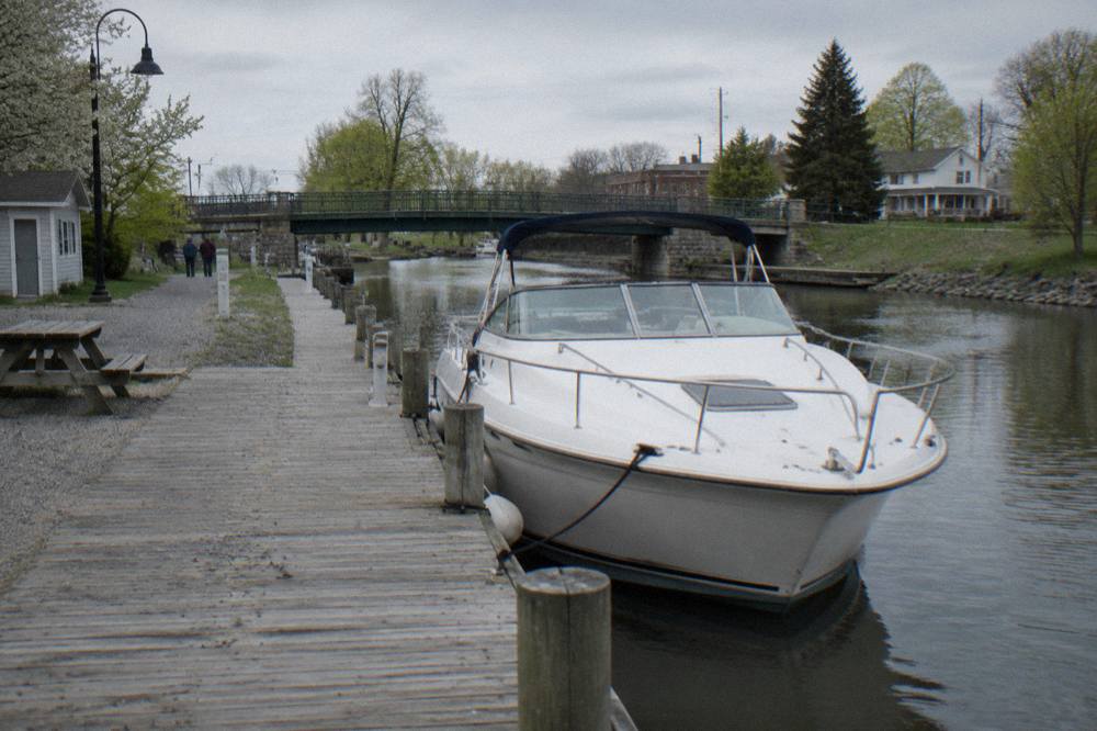 A rental canal boat moored at a dock on the Erie Canal with a village bridge in the background
