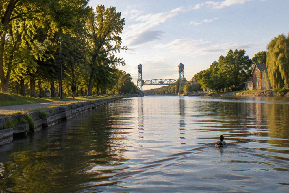 A wide view of the Erie Canal at water level with tree lined banks and a distant lift bridge