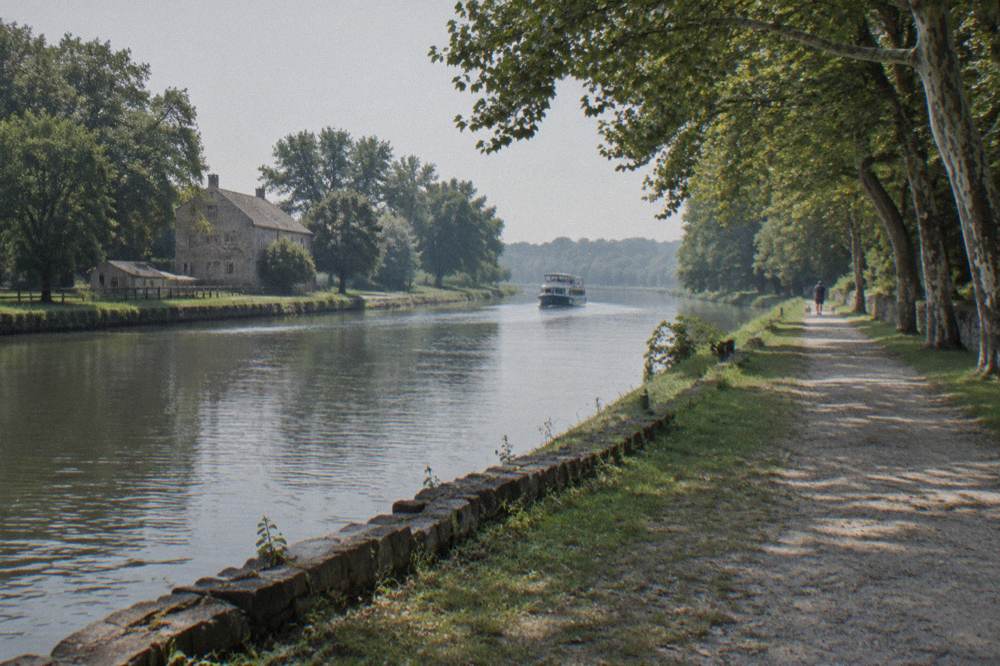 A wide view of a calm canal channel with a cruise boat in the distance and trees along the banks