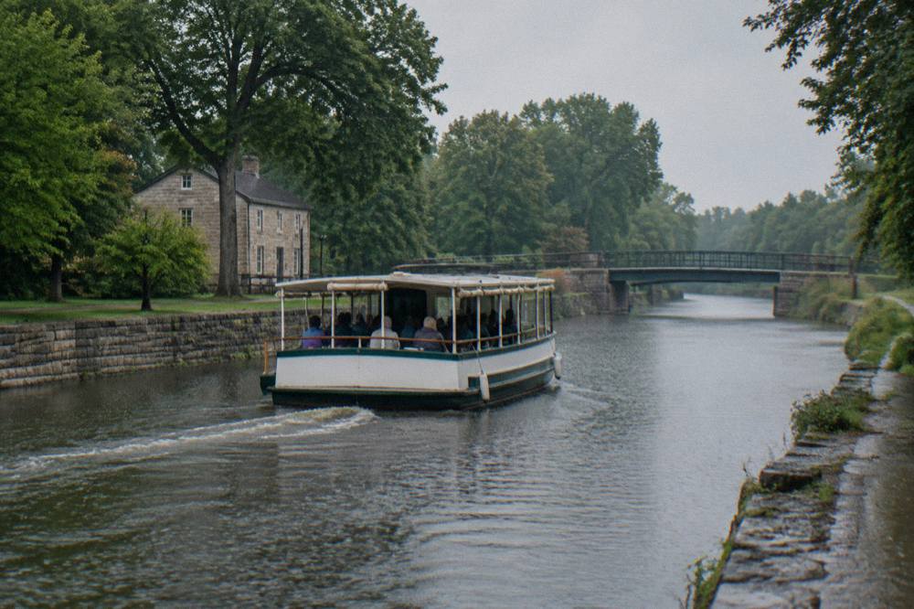 A passenger canal boat cruising a calm canal channel lined with trees and low stone embankments
