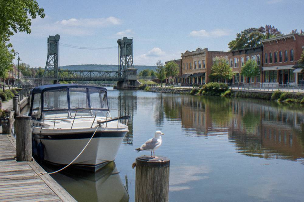 A canal boat tied at a village dock with a lift bridge and brick storefronts reflected in still canal water