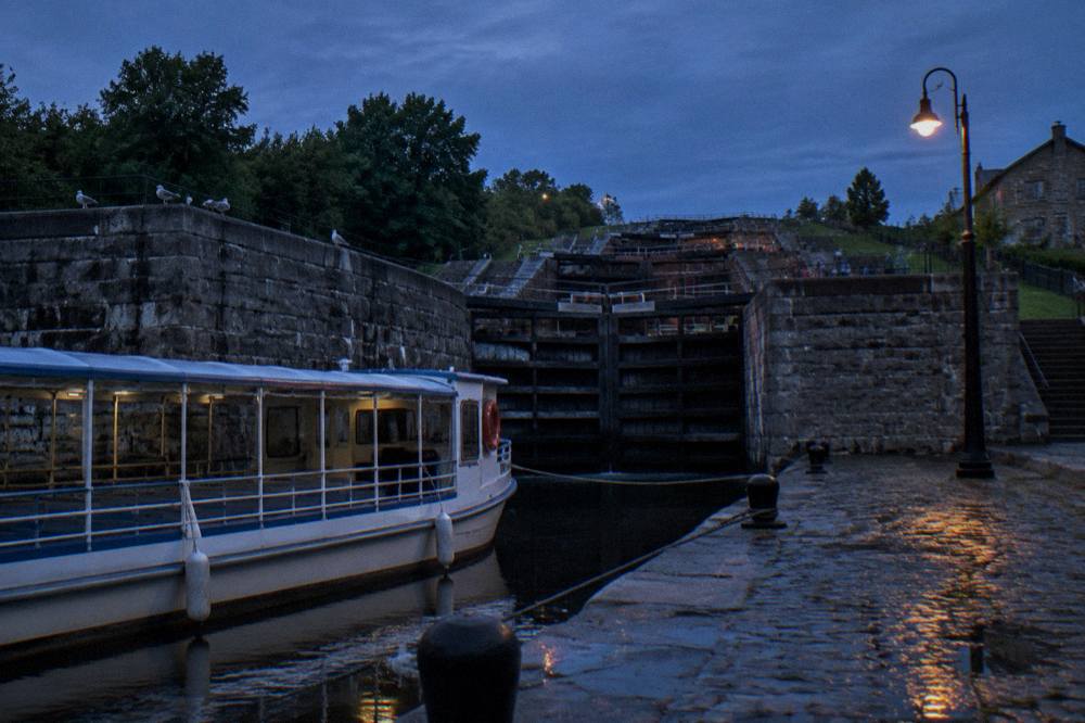 A tour boat tied at a stone dock below tall canal lock gates and a flight of older lock stonework