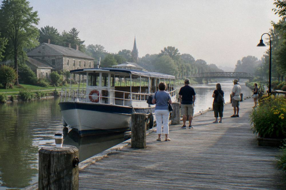 A village canal dock with a tour boat at the berth and passengers waiting to board.