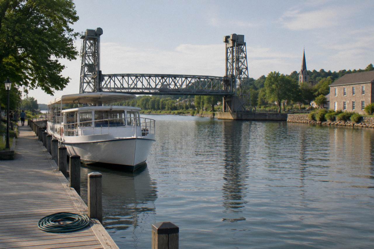 A canal tour boat tied up at a village dock beside a lift bridge on a calm afternoon.