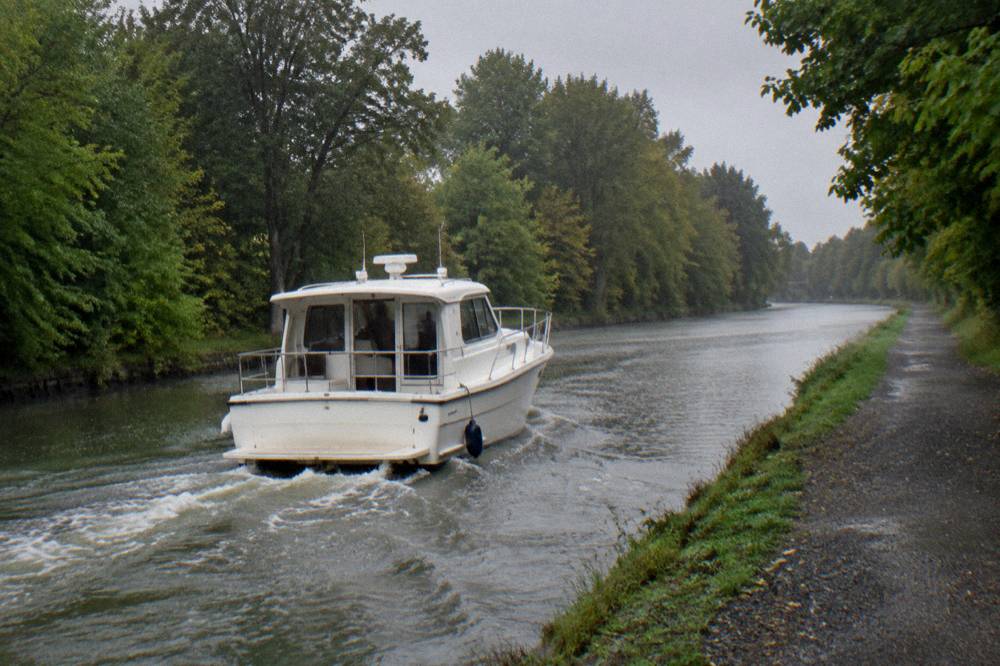 A small canal boat cruising along the Erie Canal past trees and a towpath