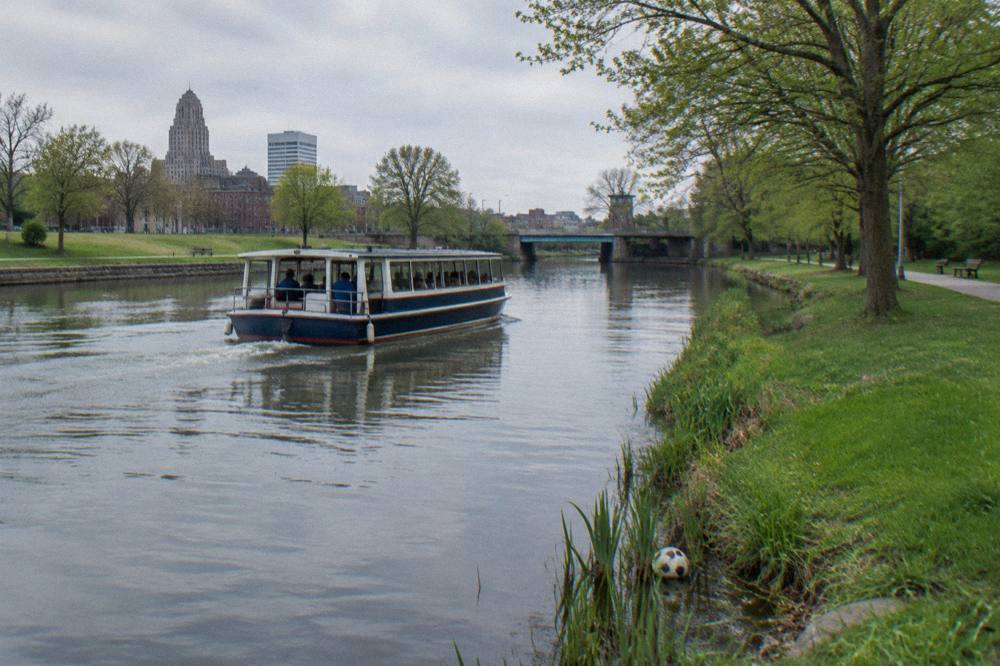 A passenger canal boat cruising past a park and a row of trees on a calm afternoon