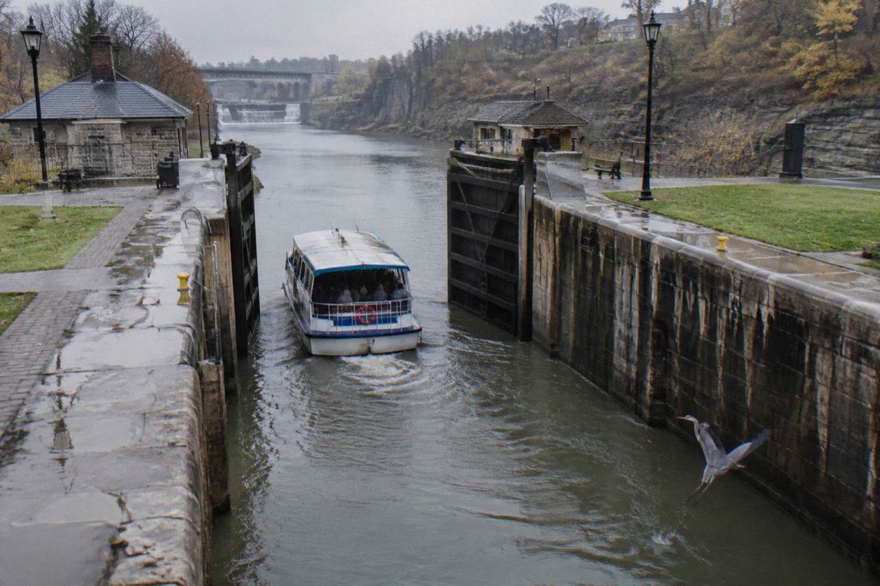 A tour boat passing through a set of Erie Canal lock gates with stone walls and a lift bridge above
