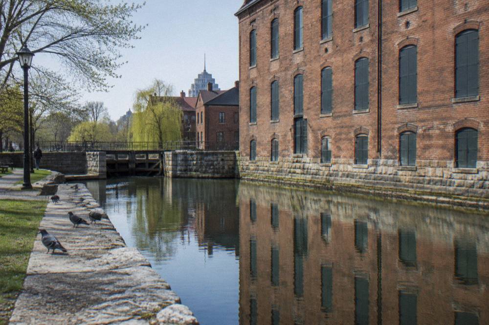 A historic brick canal-side warehouse reflected in the Erie Canal on a clear morning