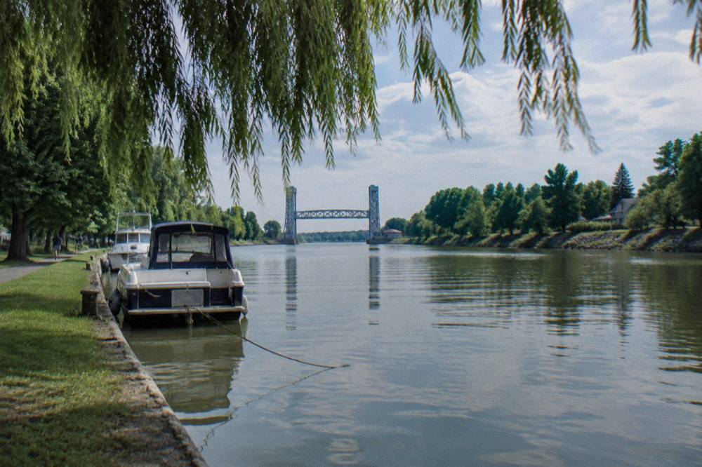 A quiet tree-lined stretch of the Erie Canal with still water and a distant lift bridge