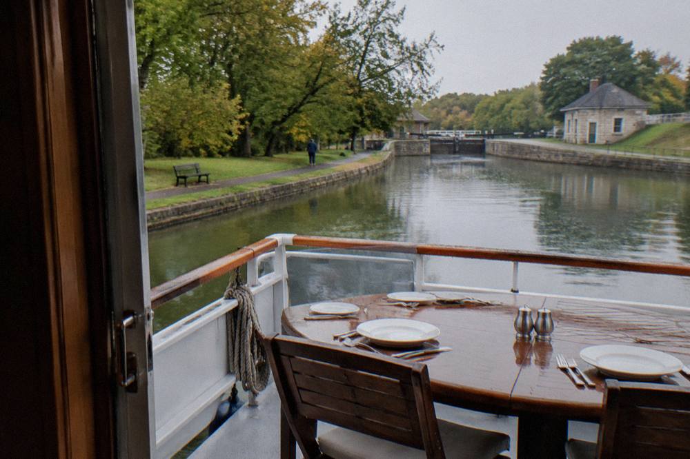 A dining table set with white plates and silverware on the deck of a canal boat, with water and trees in the background
