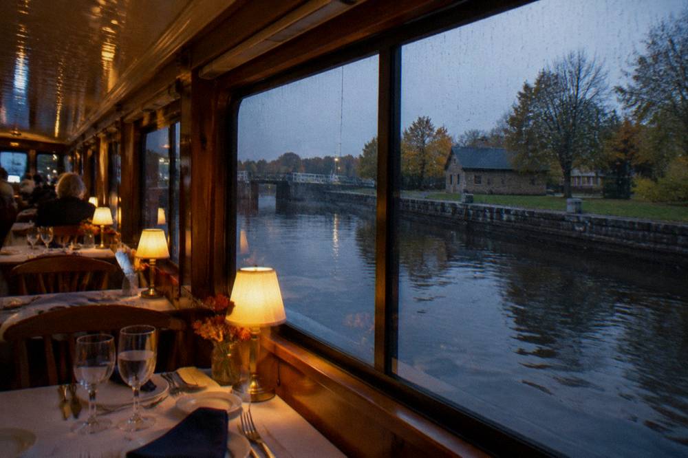 A canal boat dining area with tables set for dinner, soft lighting, and a view of the water through large windows