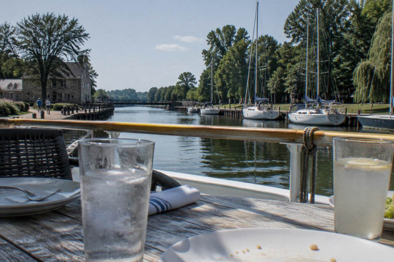 A table set with plates and glasses on the deck of a canal boat, with the Erie Canal and trees in the background.