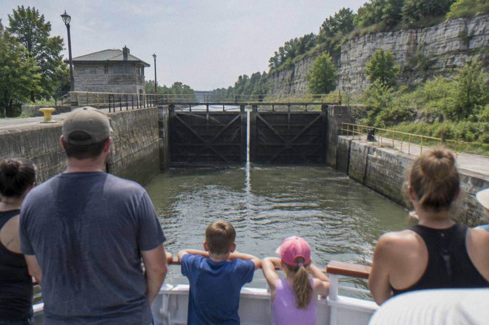 Passengers seated on the open upper deck of a canal tour boat as it approaches lock gates