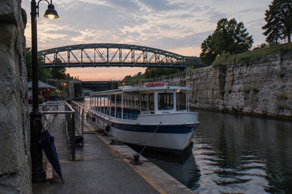A canal tour boat moored at a stone dock with lock walls and a bridge in the background