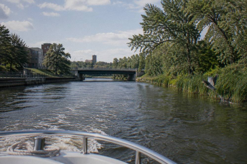 A small cruise boat moving up a wide river with tree-lined banks and a bridge ahead