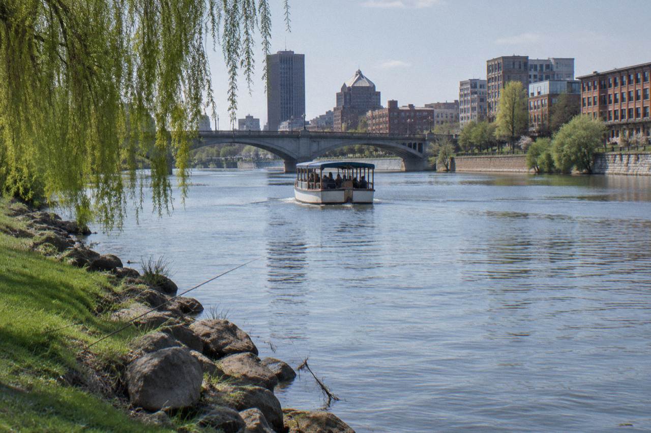 A passenger boat cruising a calm river between grassy banks and city bridges