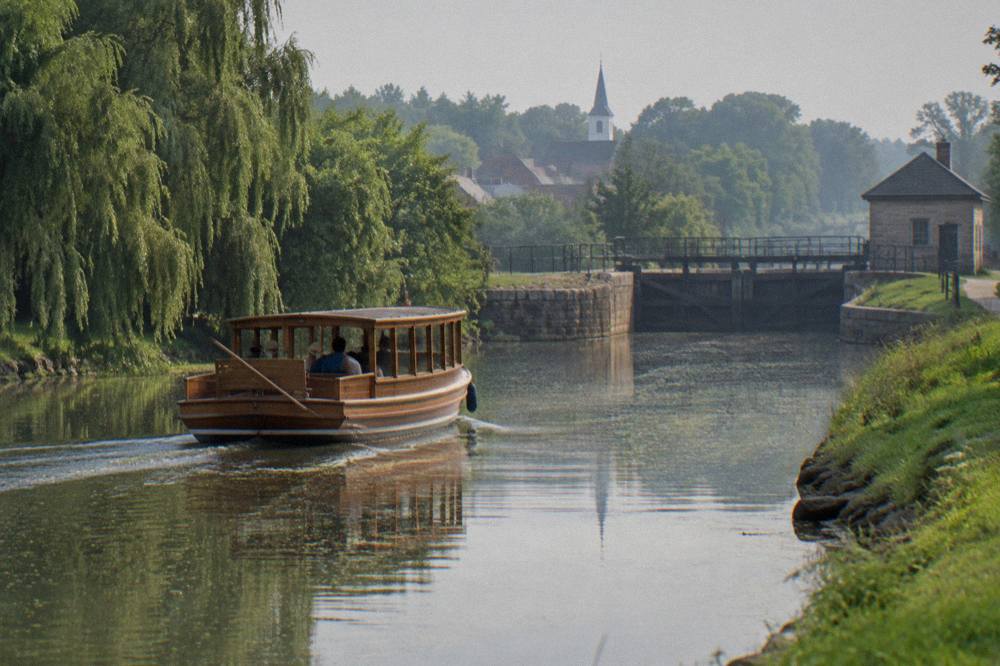 A wooden packet boat cruising past tree-lined canal banks with village buildings in the background.
