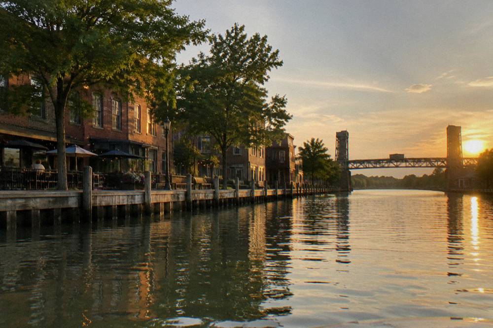 Two canal villages seen from the water, with docks and storefronts lining the canal bank under summer trees.