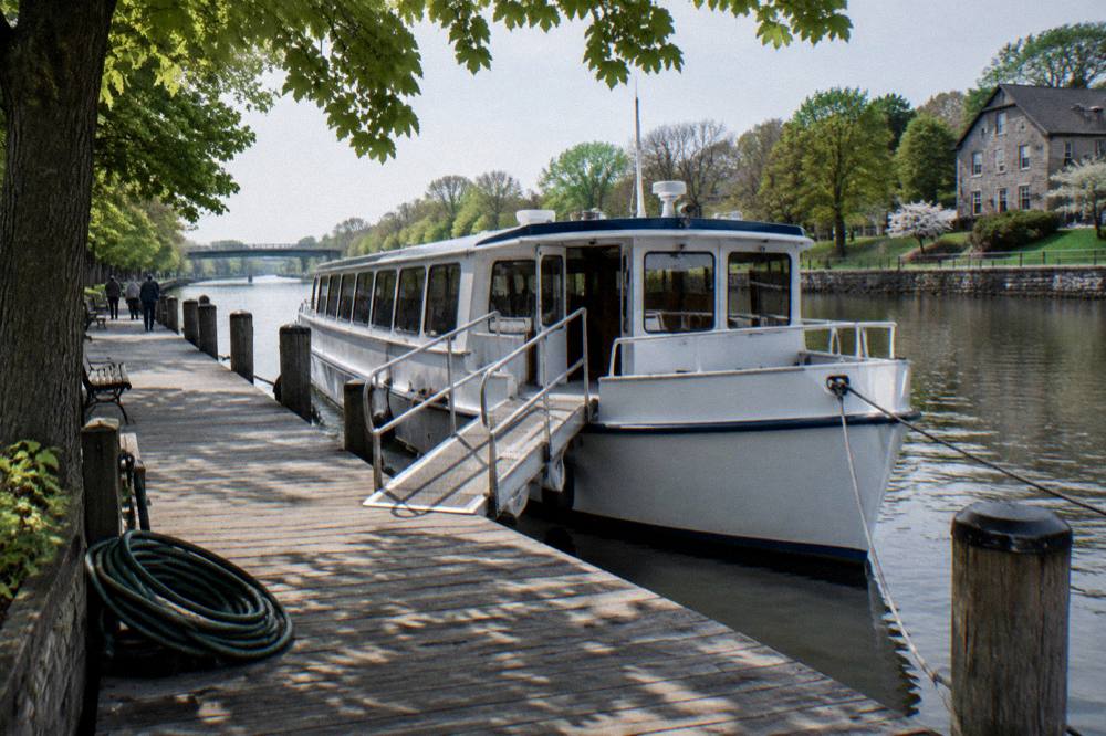 A canal boat docked at a village landing with a boarding ramp and tree-shaded walkway beside the water.