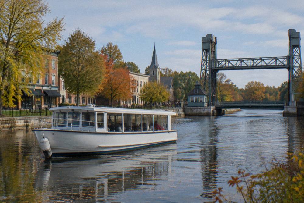 A classic canal tour boat cruising past a village main street with trees along the bank